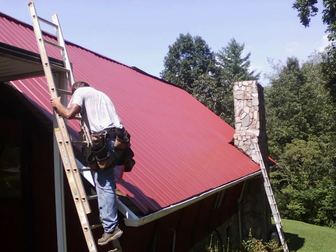 Roofer on a ladder beside a red metal roof and stone chimney — Soffit & Fascia Repair by Roofing Brothers in Pine Bush